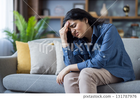 A young woman is experiencing a headache while sitting on a sofa at home, looking stressed. She is clutching her forehead. A young woman is experiencing a headache while sitting on a sofa at home, looking stressed. She is clutching her forehead. 131787976