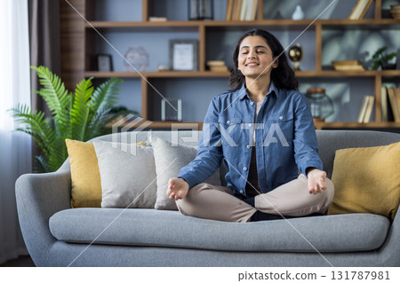 A young woman with eyes closed, meditates peacefully while sitting on a cozy sofa in her living room. The scene emphasizes calmness and well-being. 131787981