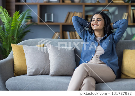 Smiling and relaxed young Indian girl sitting on the couch at home, hands behind her head and resting. 131787982