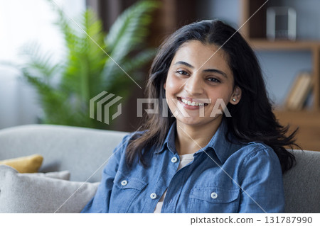 A beautiful woman with a bright smile sits comfortably on a couch. She is enjoying her time in a cozy indoor setting, indoors, and smiling. 131787990