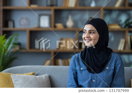 Close-up portrait of a young Muslim woman in a hijab and shirt sitting on a sofa at home, smiling and looking to the side. Close-up portrait of a young Muslim woman in a hijab and shirt sitting on a sofa at home, smiling and looking to the side. 131787999