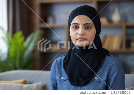 A portrait of a serene muslim woman wearing a black hijab. She is sitting inside and looking straight at the viewer. A portrait of a serene muslim woman wearing a black hijab. She is sitting inside and looking straight at the viewer. 131788001
