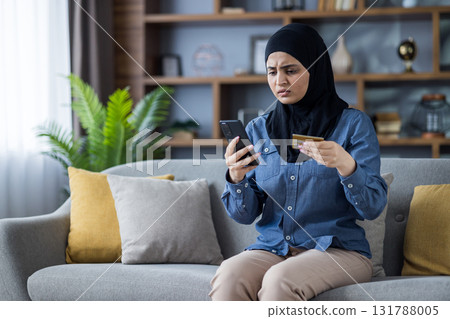 A woman wearing a hijab looks worried while holding a credit card and smartphone. She is seated on a couch. 131788005