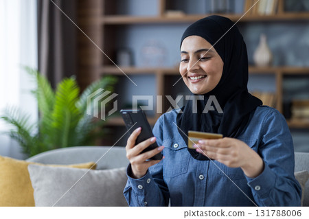 Smiling young Muslim woman wearing hijab sitting on sofa at home and using mobile phone and credit card. Smiling young Muslim woman wearing hijab sitting on sofa at home and using mobile phone and credit card. 131788006