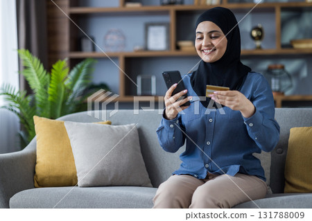 A smiling woman in a hijab makes an online purchase on her phone while holding a credit card, sitting on a sofa. 131788009