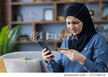 Worried Muslim young woman in hijab sitting on the sofa at home, holding a phone in her hand and looking at a credit card with sadness. 131788010
