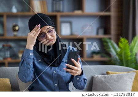 An upset Muslim woman in a hijab sits on the sofa at home and looks disappointedly at the screen ,of the phone she is holding in her hand. 131788017