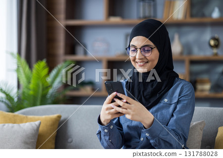Smiling young Islamic woman in hijab and glasses sitting on sofa at home and using mobile phone. Smiling young Islamic woman in hijab and glasses sitting on sofa at home and using mobile phone. 131788028