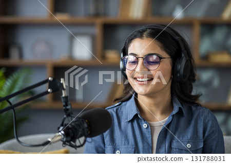 Close-up photo of a young Indian woman wearing glasses and headphones sitting at home and talking into a microphone stand. 131788031