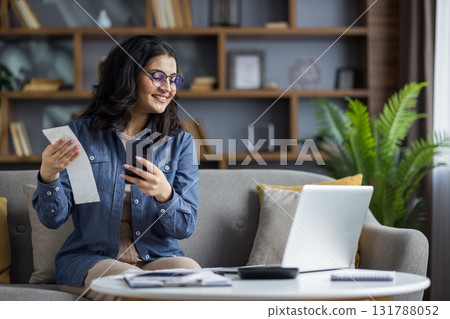 A smiling woman holds a receipt in one hand and a smartphone in the other, checking financial records. A smiling woman holds a receipt in one hand and a smartphone in the other, checking financial records. 131788052