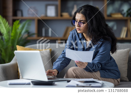 A young woman with glasses carefully reviews documents while working on her laptop at home, showing deep concentration. 131788088