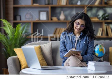 Smiling young Indian woman teaching remotely, sitting on sofa with books and laptop, taking notes in notebook. 131788183
