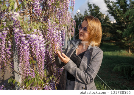 Wisteria, Woman, Garden - A woman admires purple wisteria flowers blooming on a trellis in a sunny garden. Wisteria, Woman, Garden - A woman admires purple wisteria flowers blooming on a trellis in a sunny garden. 131788595
