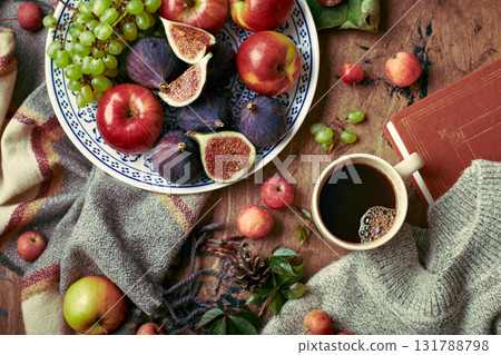 Dish with figs, apples and grapes and cup of coffee on wooden background with warm sweater, scarf, autumn leaves and apples. Autumn background, top view. Dish with figs, apples and grapes and cup of coffee on wooden background with warm sweater, scarf, autumn leaves and apples. Autumn background, top view. 131788798