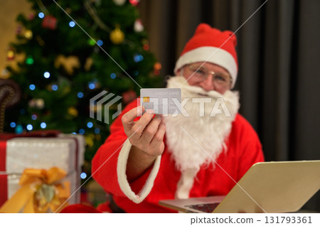 Santa claus holding credit card while using laptop for online shopping for christmas presents near decorated tree with twinkling lights in living room at home 131793361