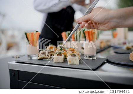 Close-up of a hand with tongs over mini rolls and vegetable sticks in cups 131794421