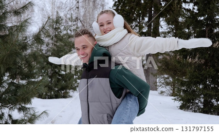 Cheerful couple enjoying winter forest, man carrying girlfriend on back, both spreading arms like wings, playful moment of connection during snowy landscape 131797155