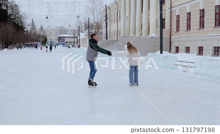 Young couple holding hands while ice skating, gliding together on snow covered rink surrounded by winter decorations in urban park setting Young couple holding hands while ice skating, gliding together on snow covered rink surrounded by winter decorations in urban park setting 131797198