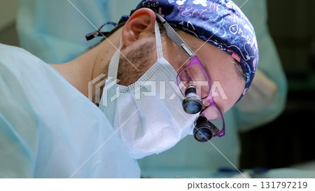 Professional surgeon wearing sterile mask, cap, glasses, surgical magnifier, carefully performing intricate medical operation in pristine operating room environment 131797219