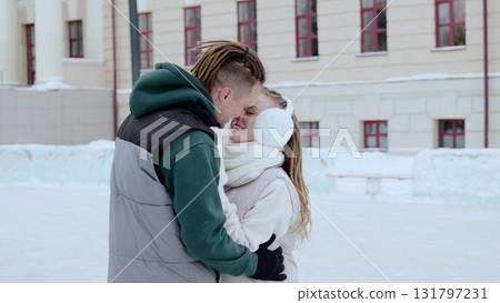 Young couple embracing, nearly kissing on winter ice rink, wearing cozy winter gear, snow covered building behind them 131797231