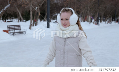 Young woman gliding gracefully on ice skates, bundled in warm earmuffs and soft scarf, radiating winter happiness in snow covered urban park landscape 131797232