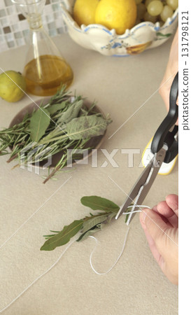 Anonymous person ties sprig of rosemary and sage together on wooden board. Spices and cooking meat at home in kitchen. 131798121