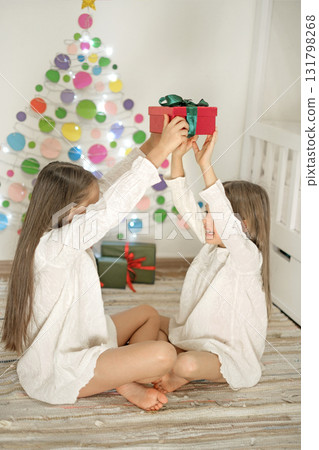 The girls are sitting on the floor of the children's bedroom, holding a Christmas present in their hands. Winter holidays. The joy of celebrating the New Year The girls are sitting on the floor of the children's bedroom, holding a Christmas present in their hands. Winter holidays. The joy of celebrating the New Year 131798268