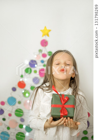 A funny little girl with a Christmas present in her hands blows a kiss. An alternative Christmas tree made of colored cardboard on the wall behind the child. A funny little girl with a Christmas present in her hands blows a kiss. An alternative Christmas tree made of colored cardboard on the wall behind the child. 131798269