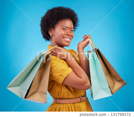 Happy black woman, shopping bag and portrait on blue background, studio and product sales. Customer, female model and smile with gift bags, retail discount promotion or luxury brand offer to consumer Happy black woman, shopping bag and portrait on blue background, studio and product sales. Customer, female model and smile with gift bags, retail discount promotion or luxury brand offer to consumer 131798572