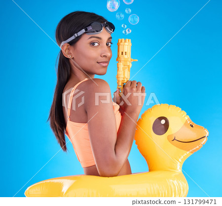 Portrait, bubbles and rubber duck with a woman on a blue background in studio ready for summer swimming. Happy, travel and vacation with an attractive or playful young female holding a bubble gun Portrait, bubbles and rubber duck with a woman on a blue background in studio ready for summer swimming. Happy, travel and vacation with an attractive or playful young female holding a bubble gun 131799471