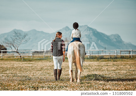 Woman leading child on horse, ranch and mountain in background lady and animal walking on field from back. Countryside lifestyle, rural nature and farm animals, mom teaching kid to ride pony in USA. Woman leading child on horse, ranch and mountain in background lady and animal walking on field from back. Countryside lifestyle, rural nature and farm animals, mom teaching kid to ride pony in USA. 131800502