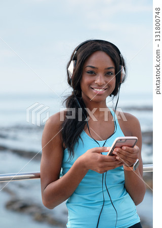 Headphones, portrait and black woman at beach with music app, fitness or sports break on smartphone. Young person on smartphone in USA on social media, networking or audio streaming technology by sea 131800748