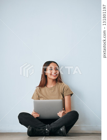 Laptop, student woman and smile by a wall with computer work for learning with happiness. University, college and house with floor sitting of a happy young person ready for studying with mockup 131801197