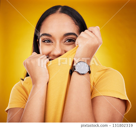 Portrait, shy and funny with a woman on a yellow background in studio pulling on her t-shirt. Face, fashion or playful and an attractive young female covering her mouth with trendy clothes for style 131802319