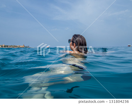 A young woman with sun glasses swimming in the blue sea. A blue hazy sky in the background 131804236