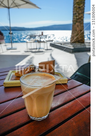 Side view of cup of coffee and croissant on wooden table by the sea. Relaxed morning breakfast scene with ocean view, sunlight, and calm atmosphere. 131806004