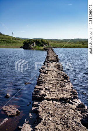 A stone path crosses a lake in Ireland, leading to a small island under a clear blue sky. A stone path crosses a lake in Ireland, leading to a small island under a clear blue sky. 131806041