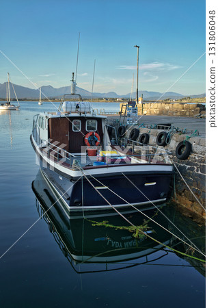 A boat is docked at a stone pier in Ireland, with mountains and other boats visible in the background under a blue sky. 131806048