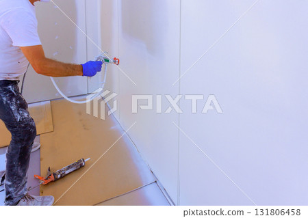Worker in gloves uses paint sprayer to coat wall with white paint at newly renovated interior space. 131806458