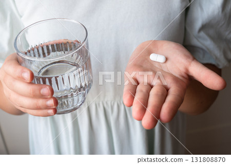 Pill and glass with water in the hand of child. Healthcare from pharmacy. Kid sit at home because of illness. 131808870