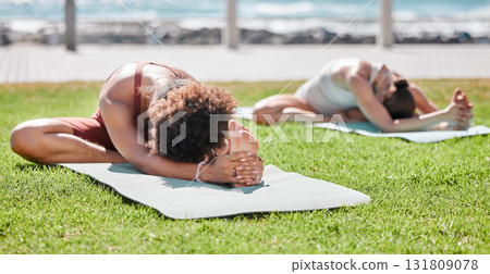 Women, yoga friends and stretching on lawn for wellness, strong legs and body goals by ocean in morning. Woman teamwork, fitness and exercise on grass with support, focus and mindfulness in summer 131809078