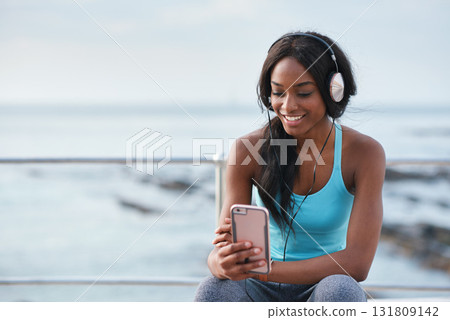 Black woman, phone and headphones at beach, sitting and smile for video, music or funny podcast to relax. Runner, rest and smartphone with meme, social media app with happiness on outdoor adventure Black woman, phone and headphones at beach, sitting and smile for video, music or funny podcast to relax. Runner, rest and smartphone with meme, social media app with happiness on outdoor adventure 131809142
