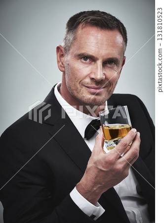 Portrait, man and a glass of whisky for celebration, smile and guy on grey studio background. Face, male and gentleman in a suit, alcohol beverage or drinking for achievement, confidence or happiness 131810523