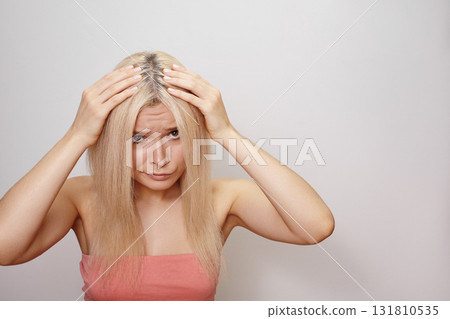 Emotive portrait of a blonde woman holding her head with both hands, expressive face and dramatic pose. Regrowth of hair roots, gray hair, hair problems, hair loss or dandruff of scalp. Beauty concept 131810535