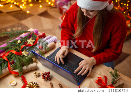 Young girl using laptop sitting at the table with many presents and decorations. Young girl ordering Christmas presents for friends and relatives, shopping in the Internet from online stores Young girl using laptop sitting at the table with many presents and decorations. Young girl ordering Christmas presents for friends and relatives, shopping in the Internet from online stores 131810754