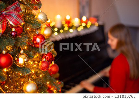 Bright decorated fir tree and young girl playing the piano on the background. Celebrating Christmas, girl playing festive New Year songs, focus on Christmas tree Bright decorated fir tree and young girl playing the piano on the background. Celebrating Christmas, girl playing festive New Year songs, focus on Christmas tree 131810765