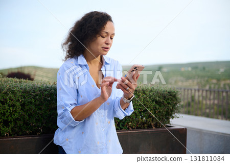 Woman Using Smartphone Outdoors By Hedge, Checking Messages On Phone During Daytime 131811084