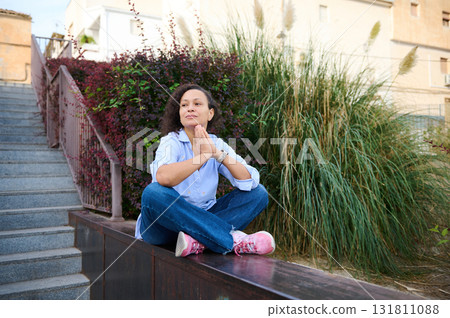 Woman Meditates Outdoors In Casual Attire On A Low Wall, Surrounded By Greenery And Stairs Woman Meditates Outdoors In Casual Attire On A Low Wall, Surrounded By Greenery And Stairs 131811088