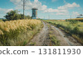 Rural dirt road lined with tall grasses leading past a water tower and fields under a blue sky 131811930