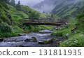 A wooden bridge spans a flowing stream amidst green hills and misty mountains 131811935